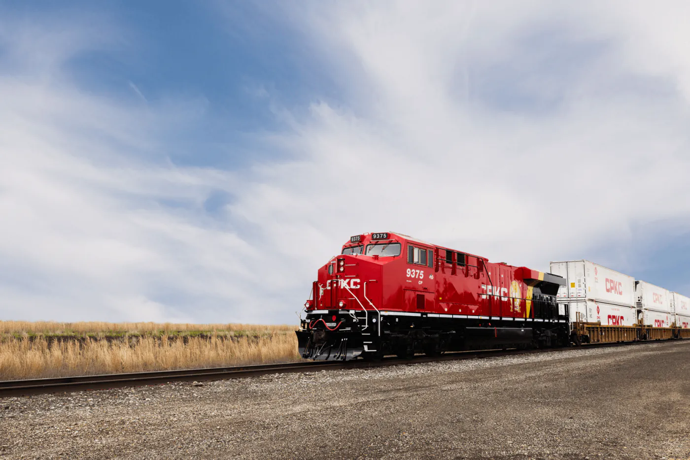 A red CPKC (Canadian Pacific Kansas City) locomotive leading a double-stacked CP intermodal container train on a single track across the open Alberta prairie under a wide blue sky