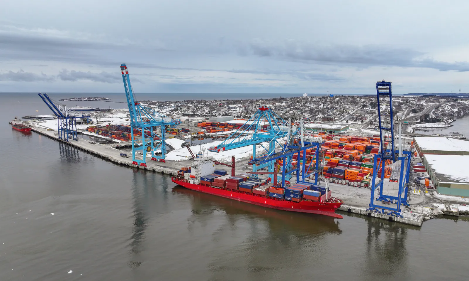 Aerial view of the DP World Saint John West Side container terminal on the Bay of Fundy — a red container ship at berth worked by blue ship-to-shore gantry cranes, with stacked containers and the snow-covered city of Saint John, New Brunswick behind
