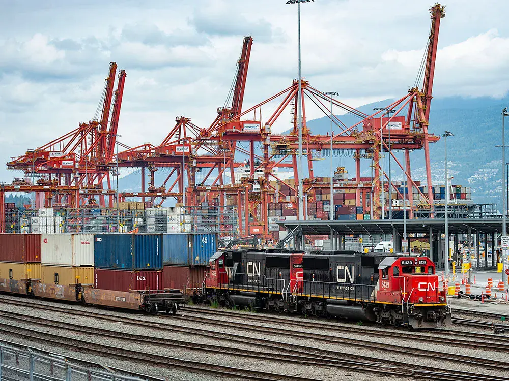CN locomotives hauling a double-stacked intermodal container train past ship-to-shore gantry cranes at a Port of Vancouver container terminal on Burrard Inlet